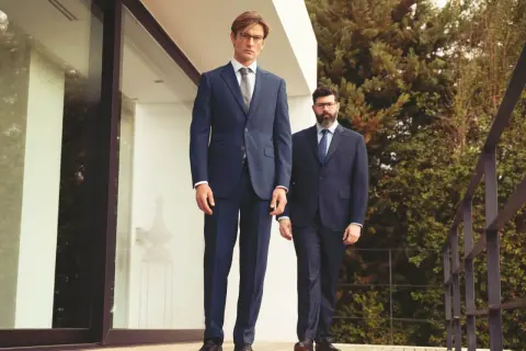 Zwei Männer in eleganten Anzügen stehen auf einer Holzterrasse. Der vordere Mann zeigt, wie man einen Anzug richtig trägt, mit perfekter Passform und Stil.
