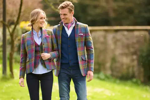 Ein Paar spaziert im Freien, beide tragen farbenfrohe Tweed-Sakkos. Sie zeigen, wie man ein Tweed-Sakko kombinieren kann, mit Westen und Jeans.
