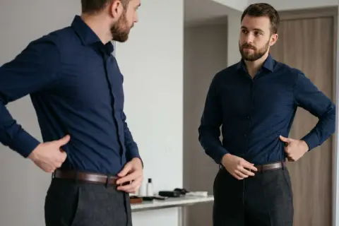 Mann im Spiegel prüft verschiedene Arten von Hemden, trägt ein dunkelblaues Hemd und graue Hose.