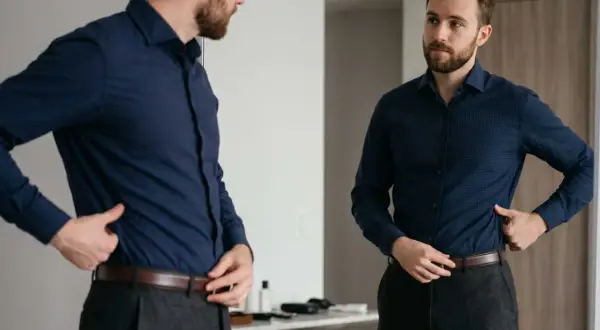 Mann im Spiegel prüft verschiedene Arten von Hemden, trägt ein dunkelblaues Hemd und graue Hose.