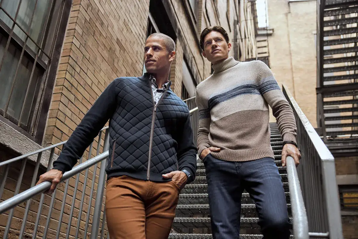 Zwei M&auml;nner auf einer Treppe, die den casual chic dress code verk&ouml;rpern. Einer tr&auml;gt eine gesteppte Jacke, der andere einen gestreiften Rollkragenpullover.
