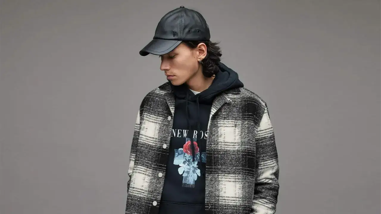 Mann mit schwarzer Lederkappe tr&auml;gt ein kariertes Hemd &uuml;ber einem Hoodie mit Rosenmotiv. Perfekt, um ein Hemd mit Hoodie zu kombinieren.
