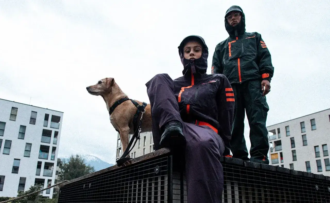 Gorpcore-Outfit mit Hund und zwei Personen in dunklen, funktionalen Jacken und Hosen vor urbaner Kulisse.