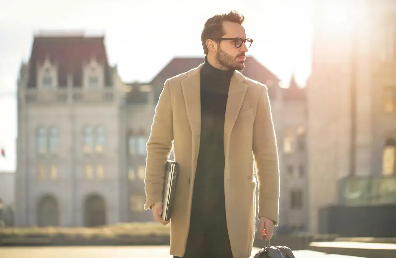 Mann in beigem Mantel und schwarzem Rollkragenpullover, der einen Laptop und eine Tasche tr&auml;gt. Stilvolle Kleidung f&uuml;r den modernen Mann.